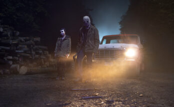Two of The Strangers standing in front of a truck in a wooded area at night. One holds an axe.